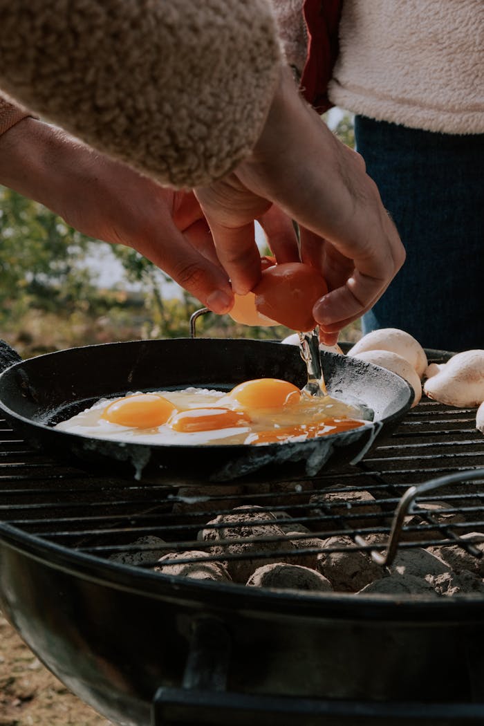 Close-up of hands cracking eggs into a frying pan on an outdoor grill.